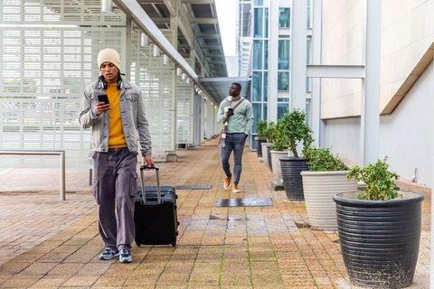 Diverse male commuters walking with rolling suitcase checking smartphones under canopy