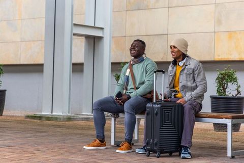 Two friends sitting on bench, waiting with suitcase and smartphone outside modern building