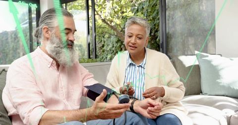 Mature couple measuring blood pressure on sofa at home with arm cuff monitor and stethoscope