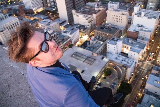 Man in blue suit and sunglasses sitting on rooftop, overlooking city skyline at dusk. Ideal for urban lifestyle, city adventures, young professionals, real estate marketing.