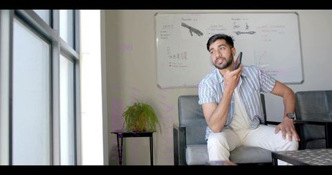 Indian man holding phone leaning and listening in office lounge with whiteboard notes