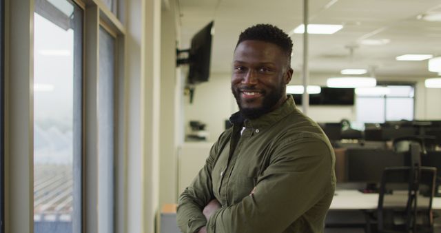 Professional man standing in modern office, smiling confidently with arms crossed near window. Ideal for business profiles, corporate websites, career promotion, and workplace diversity materials.