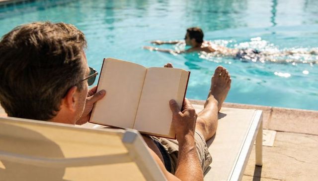 Man reading book on poolside lounger at sunlit resort pool with swimmer in background