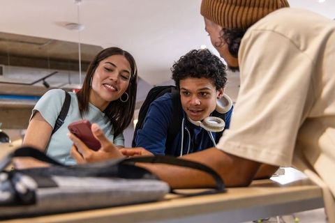 Diverse teen students collaborating over smartphone in modern university lounge with headphones