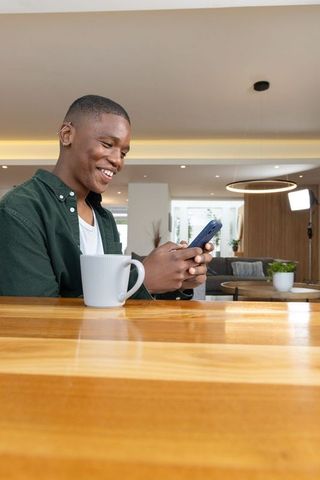 Young african american man smiling and texting on smartphone at wooden kitchen counter