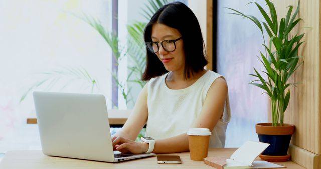 A young Asian businesswoman is working on her laptop in a bright office space, with copy space. Her focused demeanor and the presence of a smartphone and coffee cup suggest a productive work environment.