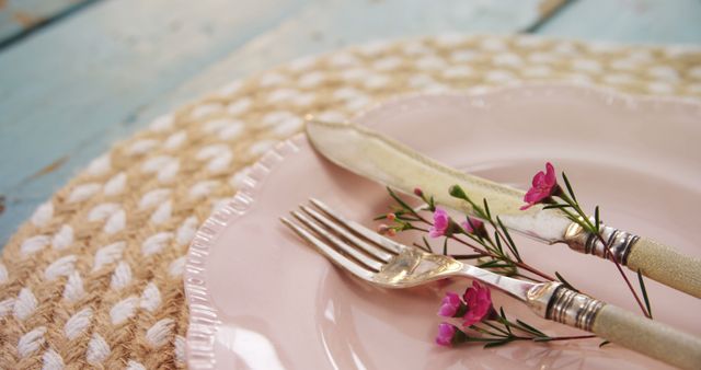 A close-up view of a table setting with a pink plate, vintage silverware, and delicate flowers, with copy space. The arrangement suggests a quaint, rustic charm, ideal for a springtime event or a romantic dinner.