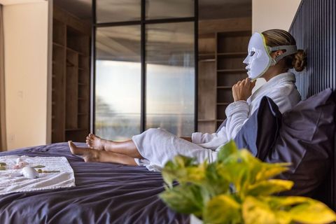 Woman wearing led facial mask relaxing on bed in modern suite for skincare and wellness
