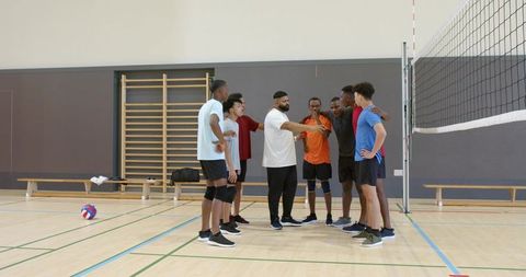 Diverse male volleyball team huddling with coach on indoor court, teamwork and leadership