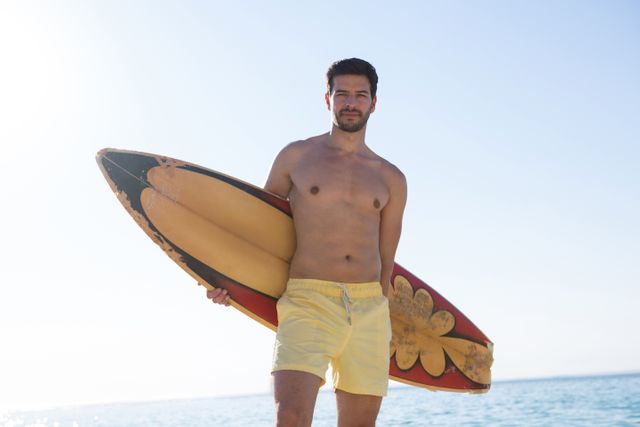 Young shirtless man holding surfboard at beach against clear blue sky on sunny day