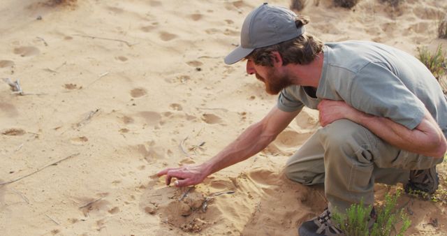 A man, wearing outdoor attire, is crouching and closely examining animal tracks in desert sand. This image can be useful for articles about wildlife tracking, outdoor adventures, desert environments, and nature research.