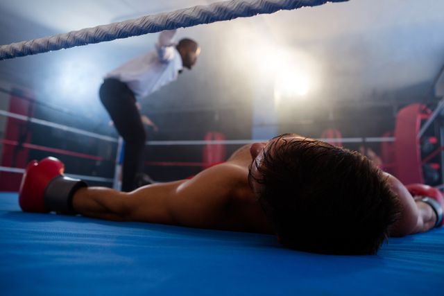 This image captures an intense moment in a boxing match where a male boxer lies unconscious on the ring floor while the referee begins the count. The scene is dramatic and filled with tension, making it suitable for use in sports-related articles, promotional materials for boxing events, fitness and training programs, or motivational content about perseverance and overcoming challenges.