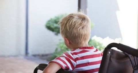 Blond toddler sitting in stroller on sunlit patio wearing red striped t-shirt looking away