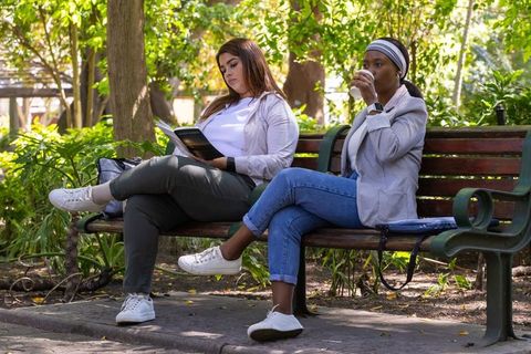 Two friends sitting on park bench reading book and sipping takeaway coffee in greenery