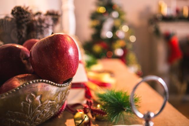 Christmas decorations with apples on a wooden table in a living room. The background features a decorated Christmas tree and festive ornaments. Ideal for holiday-themed content, home decor inspiration, and seasonal celebration promotions.