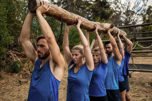 Group of fit individuals lifting a heavy wooden log together during an outdoor boot camp training session. Ideal for illustrating teamwork, physical fitness, and outdoor exercise. Useful for fitness blogs, team-building event promotions, and outdoor training programs.