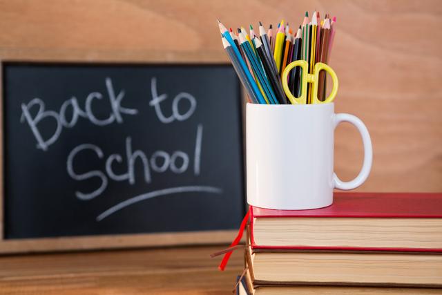 This image features a stack of books with a white mug filled with colored pencils and a pair of scissors, set against a chalkboard with 'Back to School' written on it. Ideal for educational materials, school promotions, classroom decorations, and back-to-school campaigns.