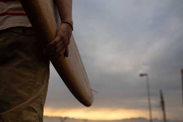 Mid section of a Caucasian male surfer wearing casual clothes and a wristband, holding a wooden surfboard and walking in an urban street at sunset.