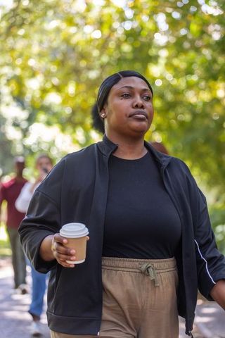 African woman walking holding coffee cup on sunlit tree-lined path for wellness