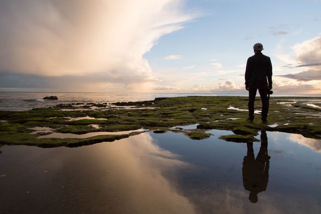 Photographer is standing on a mossy coastline during sunset, holding a camera and gazing at the horizon. Reflective pools of water mirror the dramatic sky and lighting. Can be used for travel promotions, nature and landscape themes, peaceful and contemplative settings.