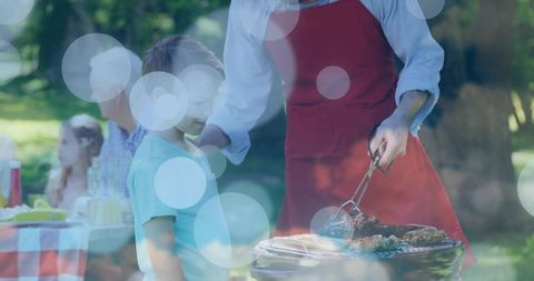Man grilling on charcoal flipping meat with tongs while child watches at family picnic
