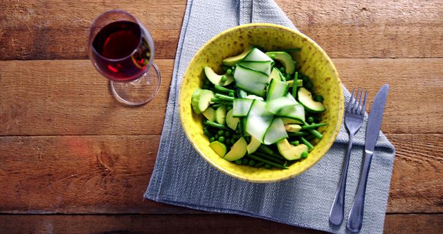 A healthy meal consisting of a green salad with sliced cucumber and avocado is served in a yellow bowl, accompanied by a glass of red wine and silverware on a wooden table. The setting suggests a focus on nutritious dining and the potential for a Mediterranean-style diet.