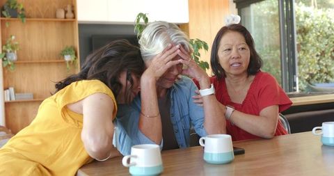 Multicultural women comforting distressed senior woman at kitchen table, sharing support