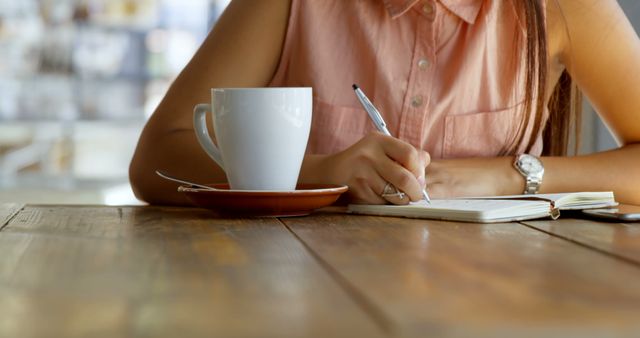 Young Asian woman writes in a notebook at a cafe. She's focused on her work, with a cup of coffee nearby, in a casual setting.