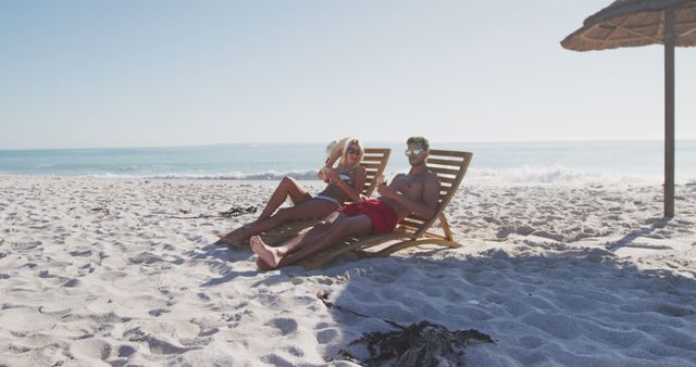 Couple relaxing on sun loungers by the beach, with a clear blue ocean and sandy shore. Ideal for travel advertisements, vacation brochures, romantic getaway promotions, summer holiday planning, leisure-themed articles, and tropical resort screenings.