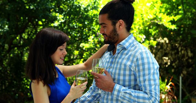 A young Asian woman and a Middle Eastern man are enjoying a toast with wine glasses in a lush garden setting, with copy space. Their cheerful interaction suggests a celebration or a romantic moment in a serene outdoor environment.