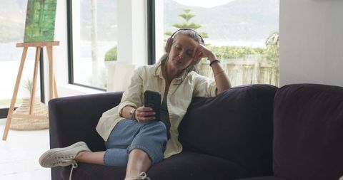 Senior woman relaxing with headphones and smartphone on modern sunlit living room sofa
