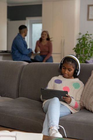 Indian girl listening on headphones using tablet on sofa while parents chat in kitchen
