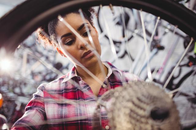 Female mechanic focusing on repairing a bicycle wheel in a workshop. She is wearing a plaid shirt and surrounded by various bike parts and tools. Ideal for use in articles or advertisements related to bicycle maintenance, professional mechanics, women in trades, and DIY repair tutorials.