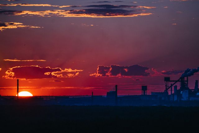 Industrial landscape featuring silhouettes of factories and smokestacks against a dramatic, red-hued sunset. Smokestacks and buildings emphasize themes of urbanization and environmental impact. Might be used for illustrations on environmental issues, urban development, industrial processes, pollution documentation, or climate change awareness campaigns.