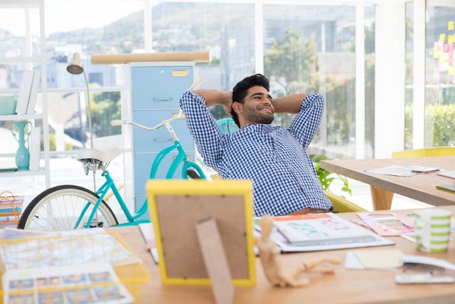 Male executive taking a break at his desk in a modern office. He is leaning back with his hands behind his head, smiling and looking relaxed. The office is bright and filled with natural light, featuring a bicycle and various office supplies. This image can be used to depict work-life balance, a relaxed work environment, or a modern office setting.
