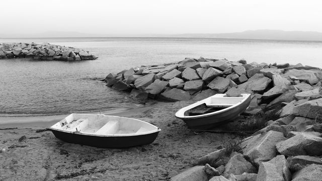 Two rowboats rest on a rocky shore by calm water in a black and white setting. This visually striking, timeless scene captures the essence of tranquility and solitude. Ideal for use in travel blogs, nautical-themed websites, or as decorative art to evoke feelings of peace and nostalgia. Could also be used in storytelling to represent journeys, quiet moments, or the simplicity of nature.