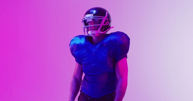 Image depicts an American football player standing under vibrant pink and purple lighting. Ideal for use in sports marketing, football gear advertisements, or promotions for athletic events. The colorful lighting makes this common subject unique and eye-catching.