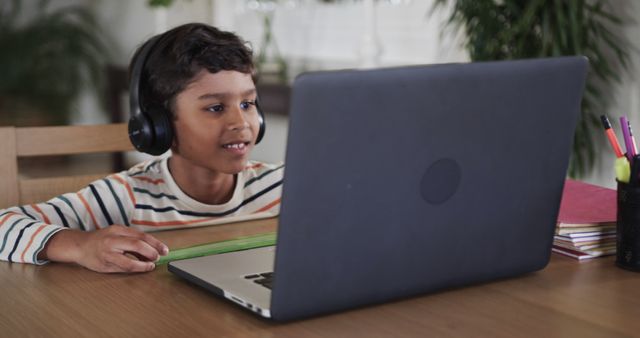 A young African American boy participates in an online lesson on a laptop while wearing headphones. This image can be used for educational technology promotions, online learning platforms, homeschooling resources, or any project highlighting children's education and distance learning.