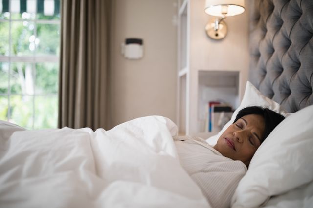 Mature woman sleeping peacefully in a cozy bedroom, wrapped in white bedding. Ideal for concepts of relaxation, rest, and comfort. Can be used in articles about sleep health, home decor, or lifestyle.