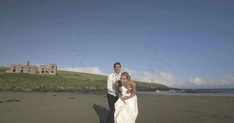 Newlyweds laughing and embracing on sandy beach by ruined castle and ocean waves