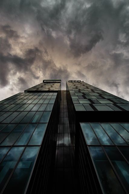 A dramatic urban scene showing a modern skyscraper with storm clouds looming overhead. The towering glass building creates a powerful contrast against the dark sky, evoking feelings of awe and tension. Ideal for use in corporate and urban-themed projects, advertisements, presentations, and media needing an intense or dramatic visual impact.