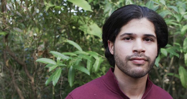 Photo captures young man with dark hair standing outdoors surrounded by green foliage. His calm and relaxed demeanor makes it suitable for use in lifestyle blogs, outdoor activities promotions, personal development materials, or nature-related content.