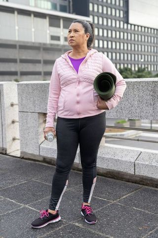 Mature woman standing holding yoga mat and water bottle on urban rooftop for fitness