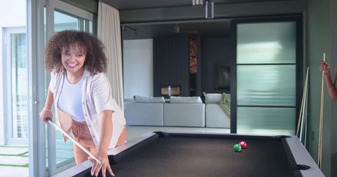 African american women playing pool in modern sunlit living room, laughing and leaning over table