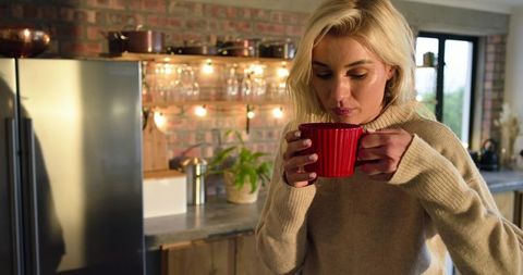 Woman holding red mug inhaling steam in cozy brick kitchen with plants and copper accents
