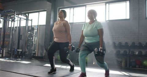 Diverse women performing weighted lunges with dumbbells in sunlit industrial gym