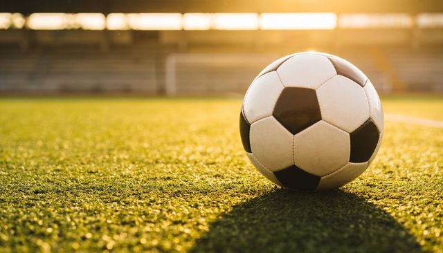 Soccer ball resting on sunlit turf at golden hour, stadium goal and long shadow