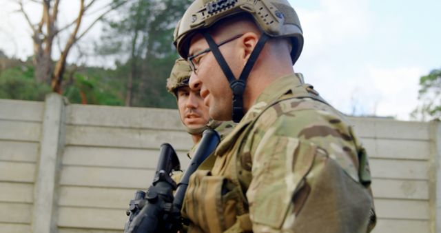 Two soldiers are outdoors wearing helmets and military uniforms discussing a strategy. This image is useful for articles or publications related to military life, teamwork and strategy planning within the armed forces, security analysis, or defense strategies.