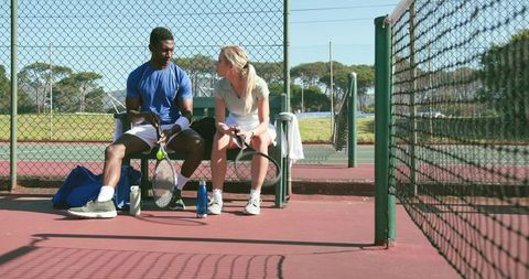 Sunlit mixed-race tennis partners sitting and talking on bench after match on outdoor hardcourt
