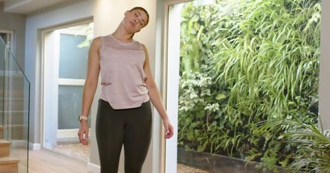 Woman stretching neck at home by glass door with garden view, smartwatch and minimalist